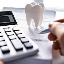 Hands holding pen and pressing calculator buttons at desk with model tooth and pile of invoices