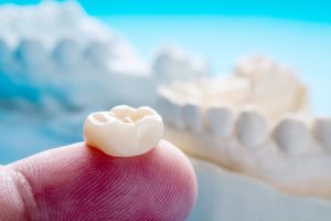 A fingertip with a dental crown placed on it in front of a blurry image of a jaw with teeth